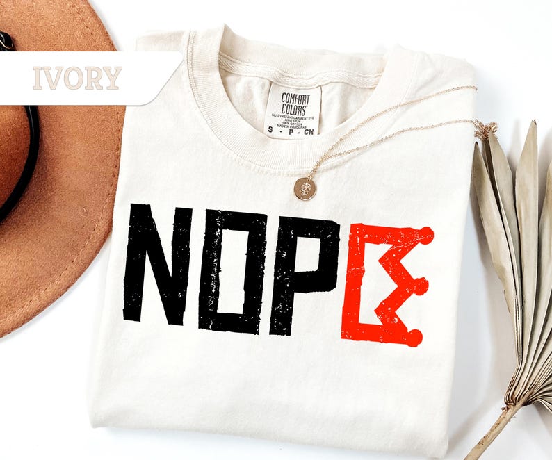 May include: Ivory-colored t-shirt with the word "NOPE" printed in black, and a red crown graphic. The shirt is folded, with a gold necklace and a hat visible in the background. The shirt is made of a soft material.