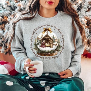 May include: A gray sweatshirt featuring a Christmas-themed graphic of a nativity scene inside a snow globe. The text around the globe reads "And you shall call His name Jesus, for He will save His people from their sins." The person is holding a white mug.
