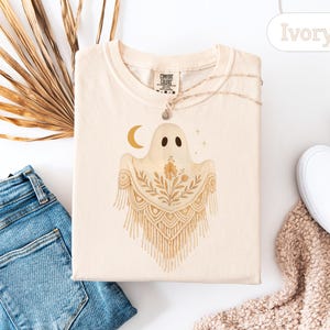 May include: Ivory-colored t-shirt featuring a whimsical ghost design with a crescent moon and floral accents. The ghost has a macrame-style shawl. The shirt is folded, with a necklace and a tag visible. The word "Ivory" is also visible.