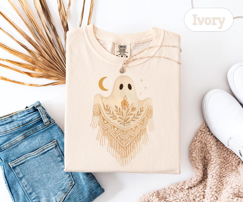 May include: Ivory-colored t-shirt featuring a whimsical ghost design with a crescent moon and floral accents. The ghost has a macrame-style shawl. The shirt is folded, with a necklace and a tag visible. The word "Ivory" is also visible.