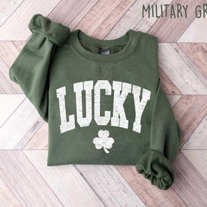 Może przedstawiać: Zielona bluza z napisem "LUCKY" w białym, przetartym stylu i białym wzorem koniczyny. Bluza jest złożona i leży na drewnianej powierzchni.