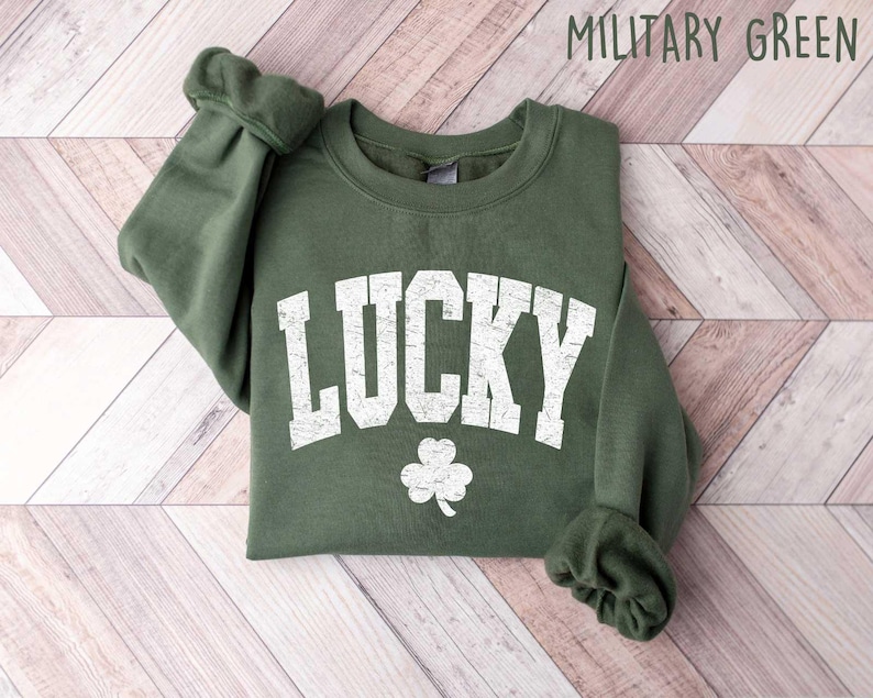 Może przedstawiać: Zielona bluza z napisem "LUCKY" w białym, przetartym stylu i białym wzorem koniczyny. Bluza jest złożona i leży na drewnianej powierzchni.