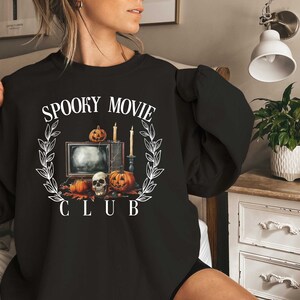 K&ouml;nnte beinhalten: Schwarzes Sweatshirt mit einem wei&szlig;en Grafikdesign eines Fernsehers, K&uuml;rbissen, Kerzen und einem Sch&auml;del mit dem Text "Spooky Movie Club".