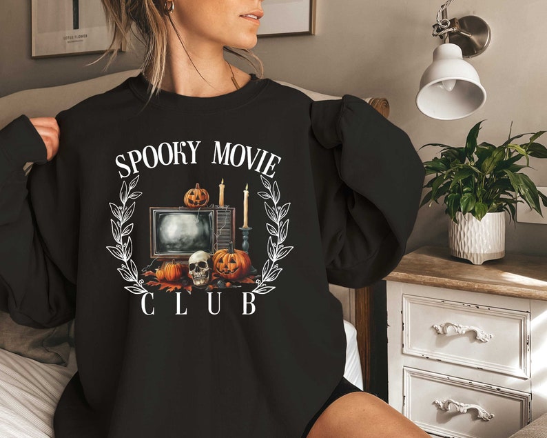 K&ouml;nnte beinhalten: Schwarzes Sweatshirt mit einem wei&szlig;en Grafikdesign eines Fernsehers, K&uuml;rbissen, Kerzen und einem Sch&auml;del mit dem Text "Spooky Movie Club".