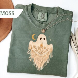 May include: Moss green Comfort Colors t-shirt featuring a ghost graphic with a crescent moon and floral accents. The ghost has a macrame-style design. A silver necklace with a pendant is on the shirt.