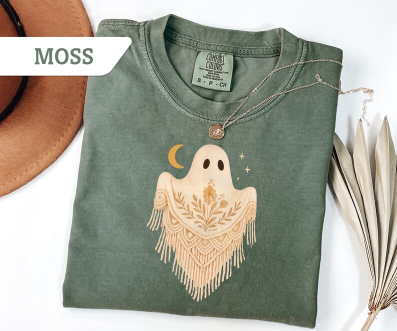 May include: Moss green Comfort Colors t-shirt featuring a ghost graphic with a crescent moon and floral accents. The ghost has a macrame-style design. A silver necklace with a pendant is on the shirt.