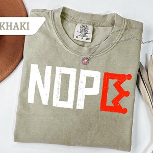 May include: Khaki-colored t-shirt with the word "NOPE" in white block letters and a red crown graphic. The shirt has a crew neck and short sleeves. A silver necklace with a pendant is draped across the shirt.