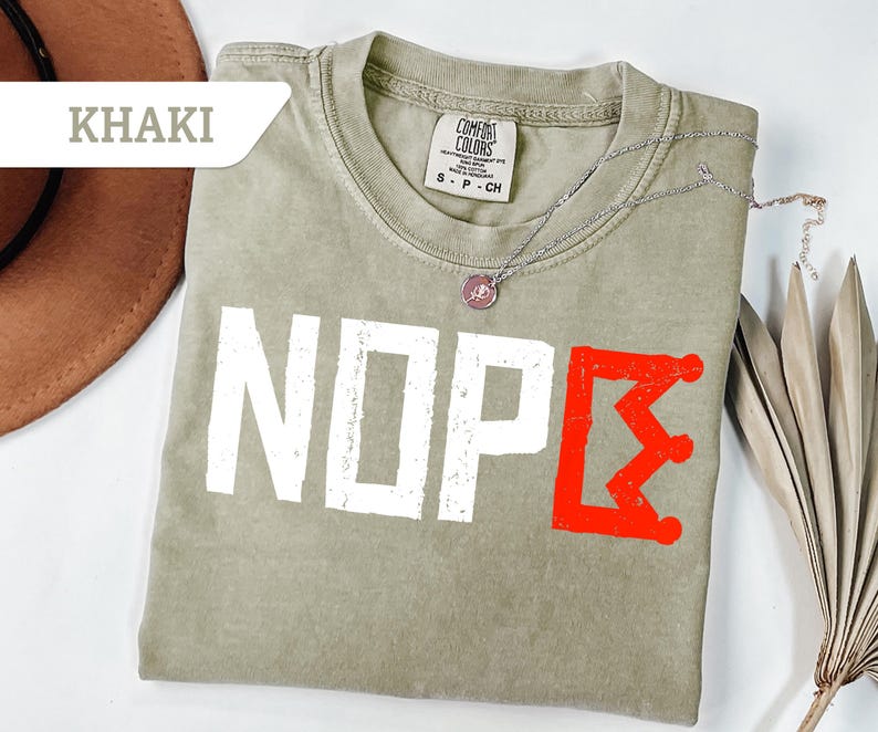 May include: Khaki-colored t-shirt with the word "NOPE" in white block letters and a red crown graphic. The shirt has a crew neck and short sleeves. A silver necklace with a pendant is draped across the shirt.