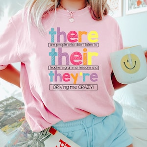Puede incluir: Una sudadera rosa claro con el texto "there are people who didn't listen to their teacher's grammar lessons, and they're driving me crazy!" en letras de colores. Se sostiene una taza blanca con una carita sonriente amarilla.