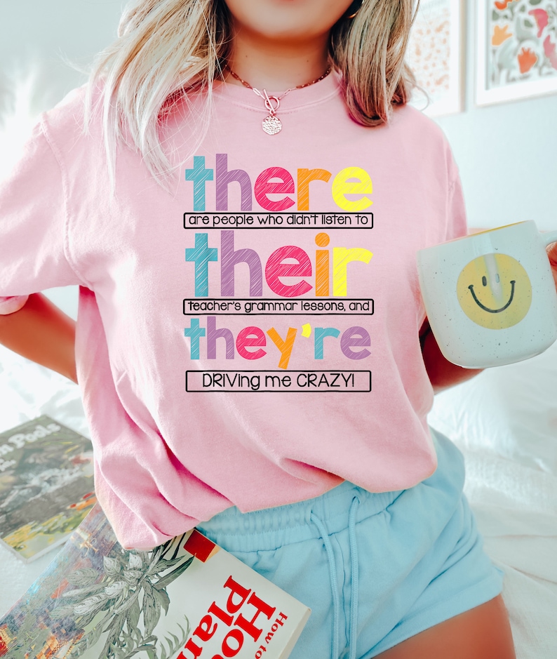 Puede incluir: Una sudadera rosa claro con el texto "there are people who didn't listen to their teacher's grammar lessons, and they're driving me crazy!" en letras de colores. Se sostiene una taza blanca con una carita sonriente amarilla.