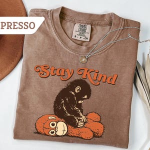 May include: Espresso-colored t-shirt with the words "Stay Kind" in orange script. A brown monkey is sitting on an orange teddy bear. A silver necklace with a small pendant is on the shirt.