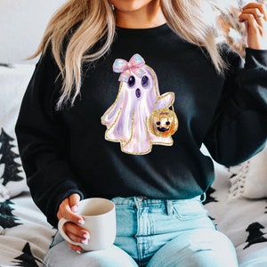 May include: Black sweatshirt featuring a watercolor ghost with a bow and a jack-o'-lantern. The ghost is white and lavender with gold glitter accents. The jack-o'-lantern is orange and gold. A fun Halloween-themed design.