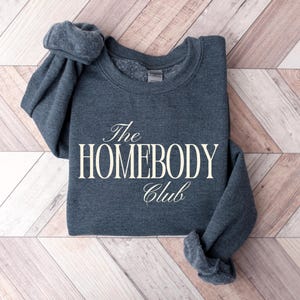 May include: A dark grey sweatshirt with the text "The HOMEBODY Club" in a white, elegant font. The sweatshirt is folded on a light brown and white wooden surface.