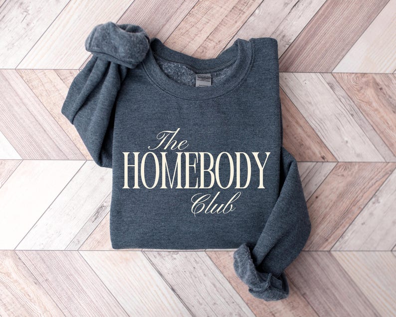 May include: A dark grey sweatshirt with the text "The HOMEBODY Club" in a white, elegant font. The sweatshirt is folded on a light brown and white wooden surface.
