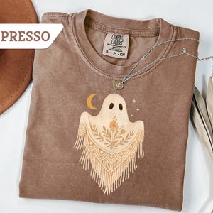May include: Espresso-colored t-shirt with a ghost graphic. The ghost has a floral design and a crescent moon. A silver necklace with a pendant is on the shirt. The shirt is a Comfort Colors brand.