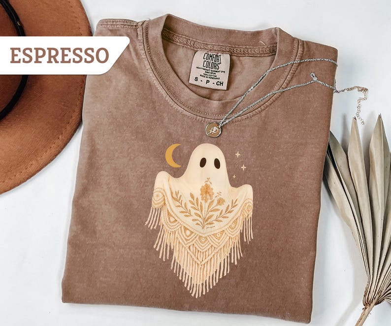 May include: Espresso-colored t-shirt with a ghost graphic. The ghost has a floral design and a crescent moon. A silver necklace with a pendant is on the shirt. The shirt is a Comfort Colors brand.