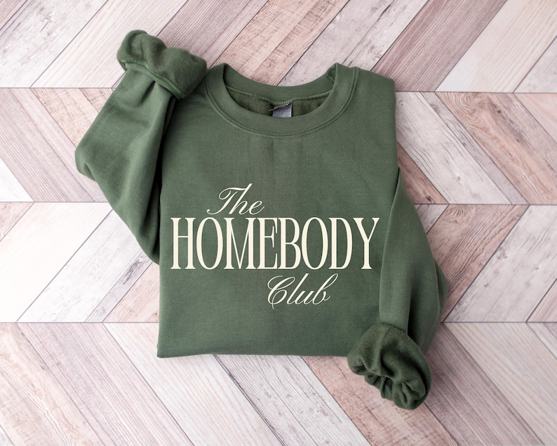 May include: An olive green sweatshirt with the text "The HOMEBODY Club" in a white, elegant font. The sweatshirt is folded neatly on a light brown and white wooden surface. The sleeves are cuffed.