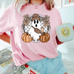 May include: A pink t-shirt with a graphic of a ghost wearing a checkered hat and holding a pumpkin and a cup of coffee. The ghost is surrounded by flowers.