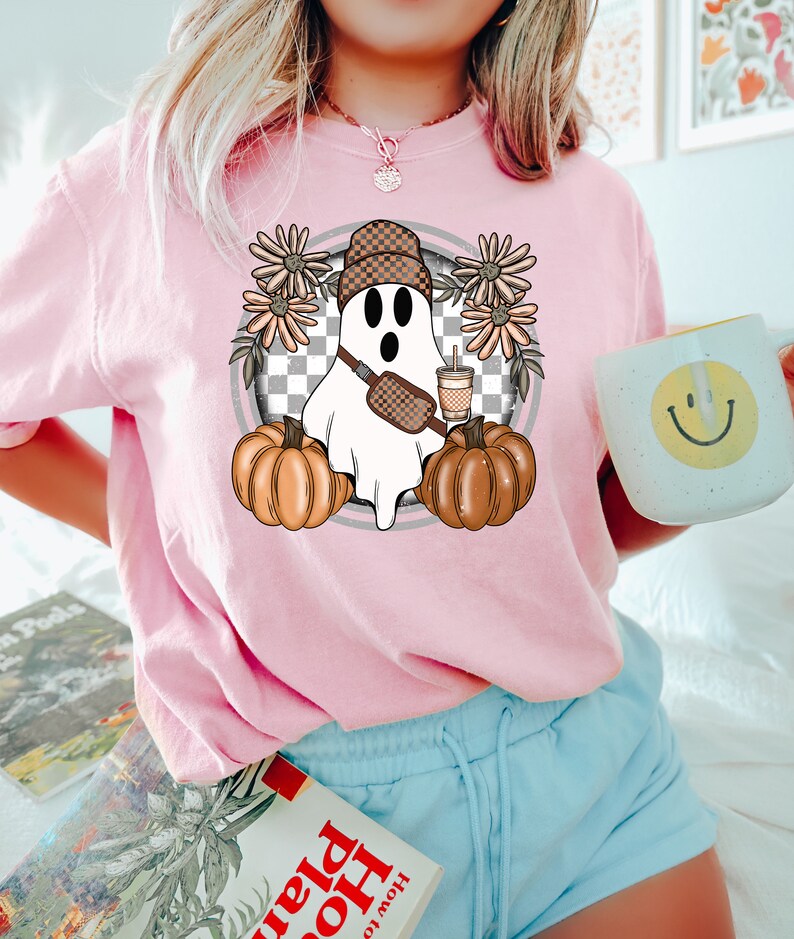 May include: A pink t-shirt with a graphic of a ghost wearing a checkered hat and holding a pumpkin and a cup of coffee. The ghost is surrounded by flowers.