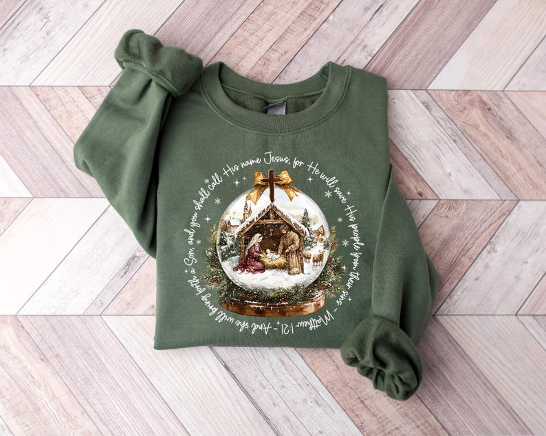 May include: Olive green sweatshirt featuring a snow globe design with a nativity scene. The snow globe has a gold bow at the top and is surrounded by text from Matthew 1:21. The sweatshirt has long sleeves and a crew neck.