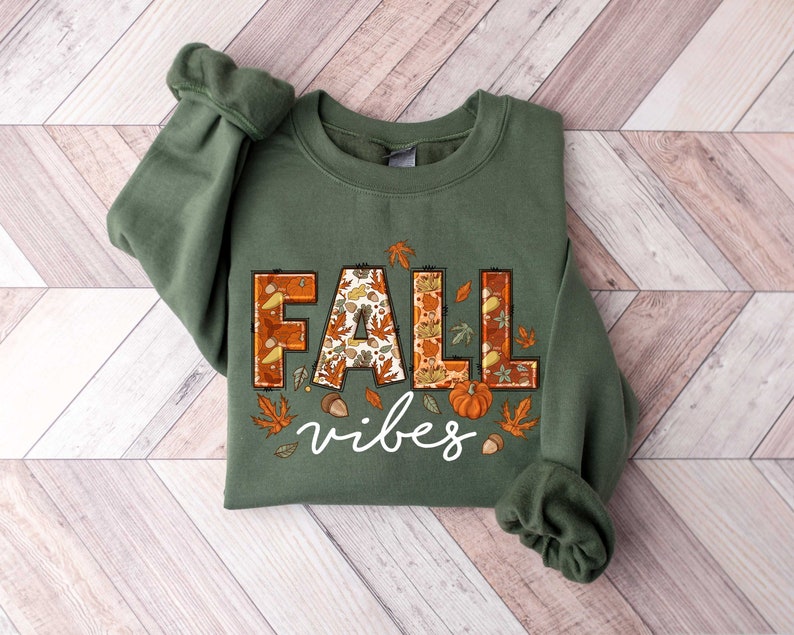 Może przedstawiać: Zielony sweter z nadrukiem "Fall vibes" z przodu. Słowo "Fall" składa się z jesiennych liści, dyni i żołędzi.