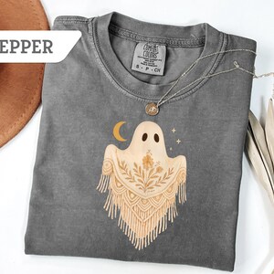 May include: A gray Comfort Colors t-shirt featuring a ghost design with a crescent moon and stars. The ghost has a floral design and fringe. The word "Pepper" is in a white banner in the upper left corner.