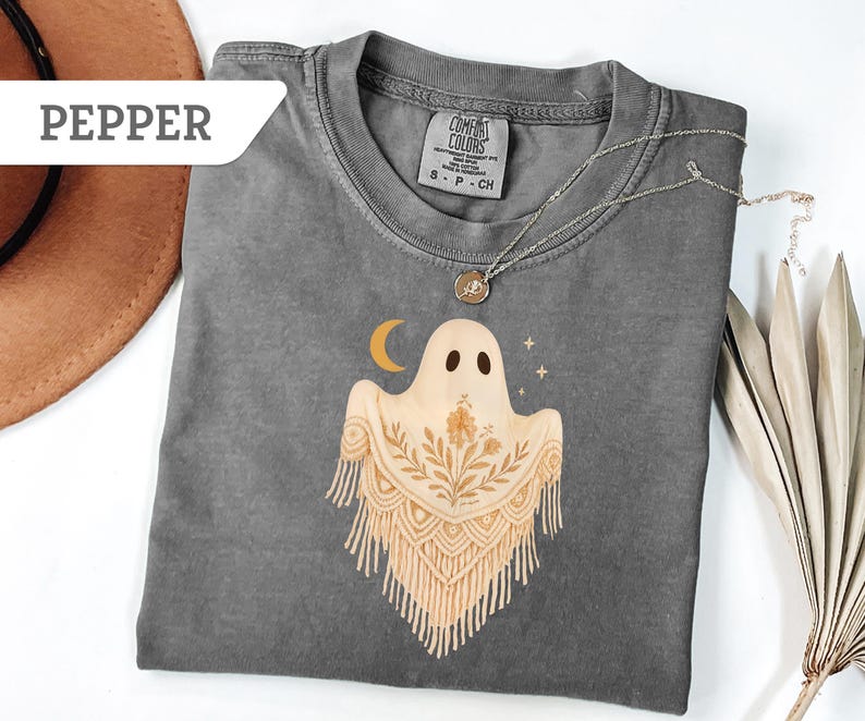 May include: A gray Comfort Colors t-shirt featuring a ghost design with a crescent moon and stars. The ghost has a floral design and fringe. The word "Pepper" is in a white banner in the upper left corner.