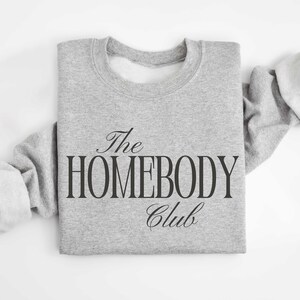 May include: A pale grey sweatshirt featuring the text "The Homebody Club" in black. The design uses a mix of script and block lettering. The sweatshirt has a crew neck and long sleeves. The image is taken from above.