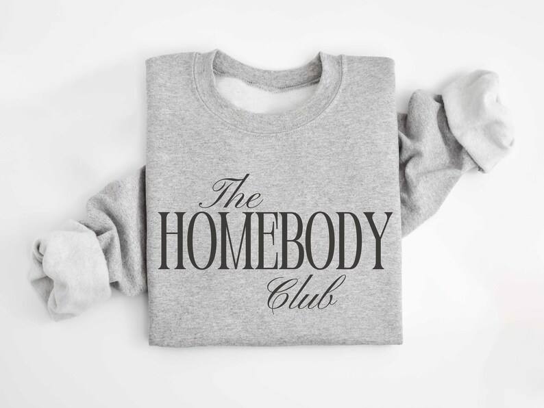 May include: A pale grey sweatshirt featuring the text "The Homebody Club" in black. The design uses a mix of script and block lettering. The sweatshirt has a crew neck and long sleeves. The image is taken from above.