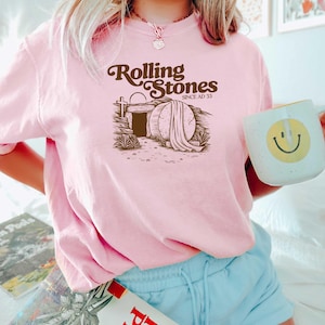 May include: Pink t-shirt with a brown graphic of a tomb with the text "Rolling Stones Since AD 33".