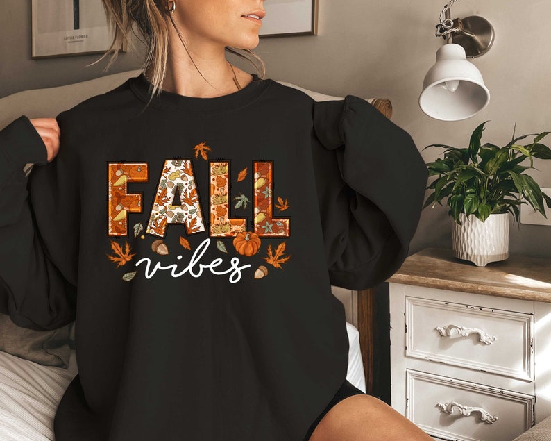 Może przedstawiać: Czarny sweter z nadrukiem graficznym słowa "FALL" w kolorze pomarańczowym i brązowym z jesiennymi liśćmi i dyniami. Tekst "vibes" jest nadrukowany pod słowem "FALL".