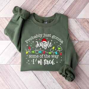 Peut inclure: Un sweat-shirt vert olive avec le texte "Probably just gonna jingle some of the way I'm tired." Le motif comprend un bonnet de P&egrave;re No&euml;l, des guirlandes lumineuses et des flocons de neige. Le sweat-shirt est pli&eacute; sur une surface en bois clair.