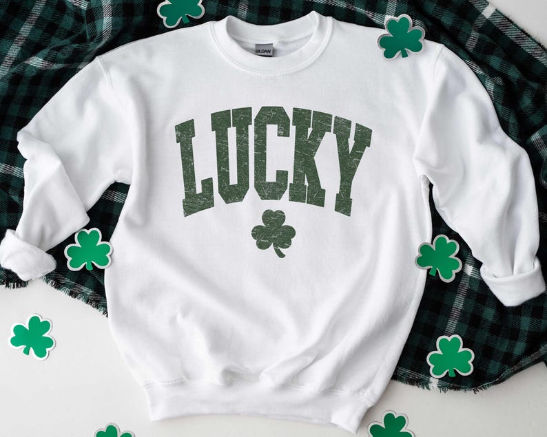 Może przedstawiać: Biała bluza z nadrukiem słowa "LUCKY" w kolorze zielonym na przodzie. Słowo jest w stylu vintage. Pod słowem znajduje się zielona koniczyna.