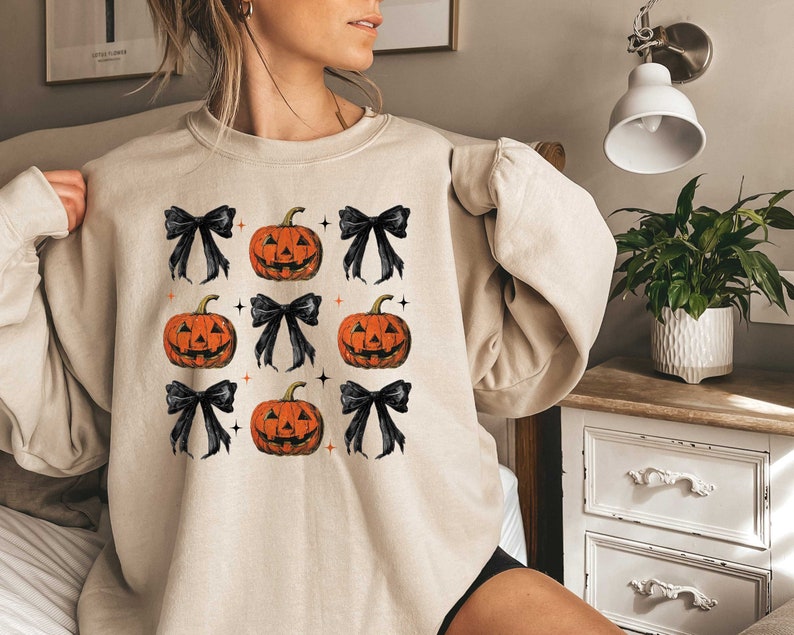 Op de afbeelding: Een beige sweatshirt met een patroon van zwarte strikken en oranje pompoenen. De pompoenen hebben een jack-o'-lantern gezicht.