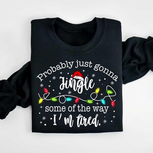 Peut inclure: Sweat-shirt noir avec le texte "Probably just gonna jingle some of the way, I'm tired." Le mot "Jingle" est surmont&eacute; d'un bonnet de P&egrave;re No&euml;l, et le motif comprend des guirlandes lumineuses et des flocons de neige.