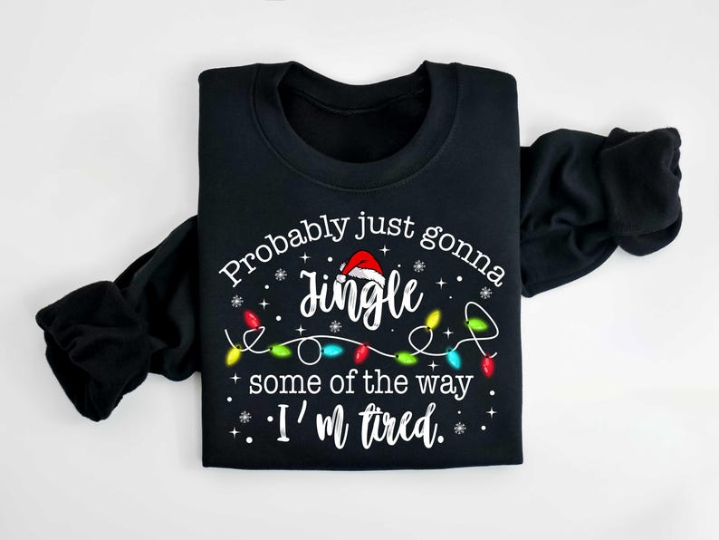 Peut inclure: Sweat-shirt noir avec le texte "Probably just gonna jingle some of the way, I'm tired." Le mot "Jingle" est surmont&eacute; d'un bonnet de P&egrave;re No&euml;l, et le motif comprend des guirlandes lumineuses et des flocons de neige.