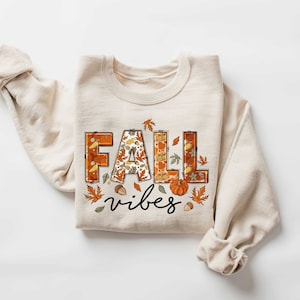 Może przedstawiać: Beżowa bluza z nadrukiem "FALL vibes" z przodu. Litery są ozdobione jesiennym wzorem z liści, dyni i żołędzi.