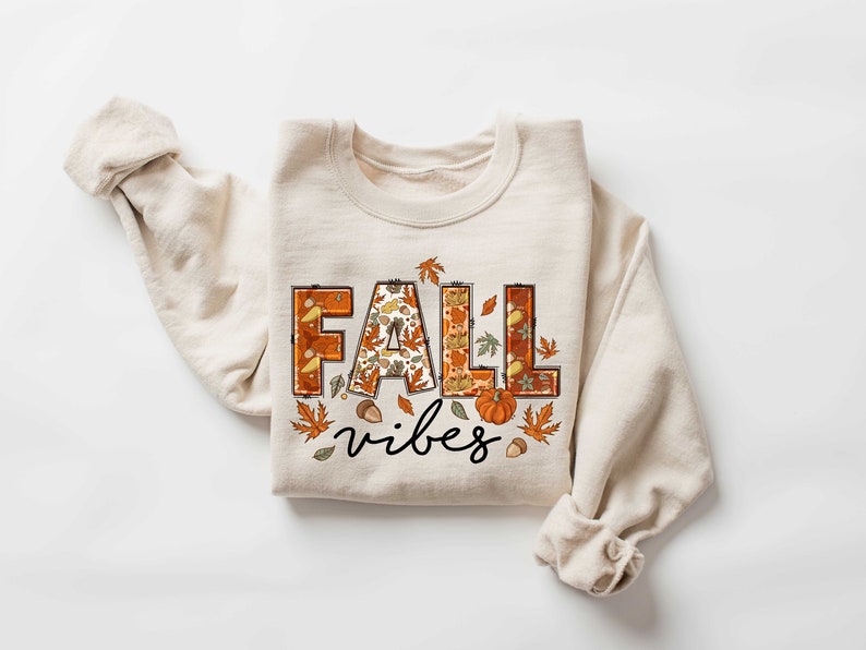 Może przedstawiać: Beżowa bluza z nadrukiem "FALL vibes" z przodu. Litery są ozdobione jesiennym wzorem z liści, dyni i żołędzi.
