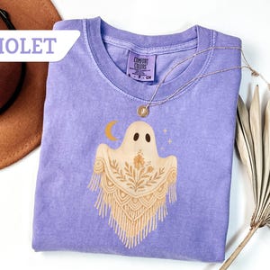 May include: Violet Comfort Colors t-shirt featuring a ghost design with a crescent moon and stars. The ghost has a floral shawl. A gold necklace is draped on the shirt. The word "VIOLET" is in a white banner in the upper left corner.