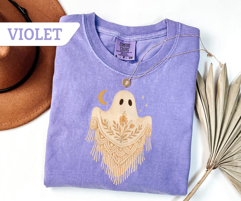 May include: Violet Comfort Colors t-shirt featuring a ghost design with a crescent moon and stars. The ghost has a floral shawl. A gold necklace is draped on the shirt. The word "VIOLET" is in a white banner in the upper left corner.