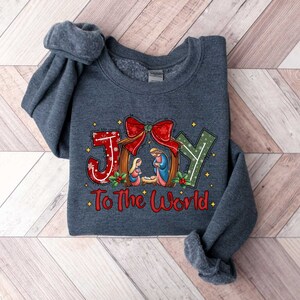May include: A dark grey sweatshirt with a Christmas-themed design. The design features the word "JOY" in red, white and green with a red bow and a nativity scene. The text "To The World" is written below the word "JOY".