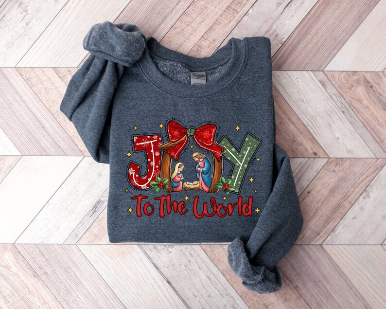 May include: A dark grey sweatshirt with a Christmas-themed design. The design features the word "JOY" in red, white and green with a red bow and a nativity scene. The text "To The World" is written below the word "JOY".