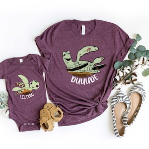May include: Two purple t-shirts featuring a cartoon sea turtle design. The larger shirt reads "DUDE" and the smaller shirt reads "LIL DUDE".