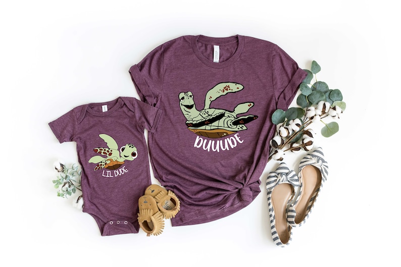 May include: Two purple t-shirts featuring a cartoon sea turtle design. The larger shirt reads "DUDE" and the smaller shirt reads "LIL DUDE".