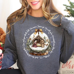 May include: A dark gray sweatshirt featuring a Christmas snow globe design with a nativity scene inside. The text around the globe reads "And you shall call His name Jesus, for He will save His people from their sins - Matthew 1:21".