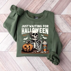 May include: Green sweatshirt with a skeleton holding a pumpkin and a coffee cup. The text "Just Waiting for Halloween" is printed on the front.