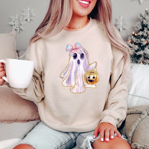 May include: Beige sweatshirt featuring a watercolor ghost illustration with a bow and a pumpkin bucket. The ghost has a gold glitter outline. The sweatshirt is a neutral color, suitable for fall and Halloween.