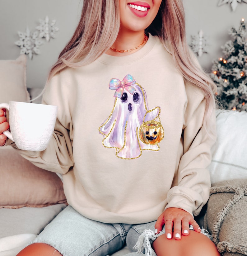May include: Beige sweatshirt featuring a watercolor ghost illustration with a bow and a pumpkin bucket. The ghost has a gold glitter outline. The sweatshirt is a neutral color, suitable for fall and Halloween.