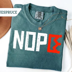 May include: A teal Comfort Colors t-shirt with the word "NOPE" in white block letters and a red Greek letter. A gold necklace with a pendant rests on the shirt. A brown hat and dried plant are in the background.