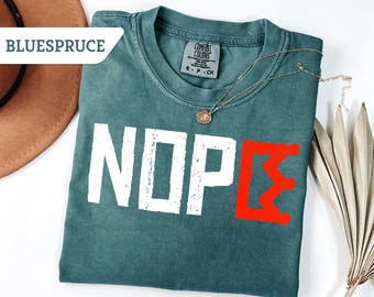 May include: A teal Comfort Colors t-shirt with the word "NOPE" in white block letters and a red Greek letter. A gold necklace with a pendant rests on the shirt. A brown hat and dried plant are in the background.